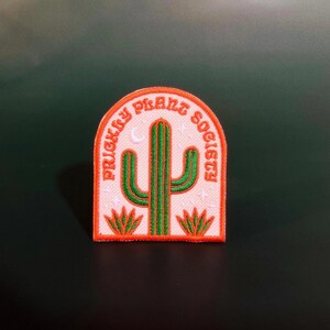 May include: Embroidered patch with the text "Prickly Plant Society" in orange, arched over a green cactus design. The patch has an off-white background and orange border, with small star and moon details.
