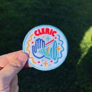 May include: Embroidered patch with the word "CLERIC" in red, arched across the top. The patch features a blue hand, a blue triangle, and other colorful accents on a white background.