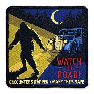 May include: Embroidered patch featuring a Bigfoot silhouette walking towards a vintage blue truck under a crescent moon. The patch has the text "WATCH THE ROAD!" and "ENCOUNTERS HAPPEN • MAKE THEM SAFE" in red and yellow.
