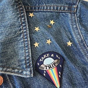 May include: A denim jacket with gold star embellishments and a patch that reads "TAKE A TRIP." The patch features a UFO design with a rainbow trail. The jacket has a classic collar and button closure.