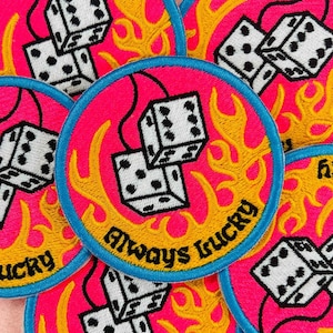 May include: Round patch with a pink background, blue border, and yellow flames. The patch features two white dice with black dots and the text "Always Lucky" in yellow.