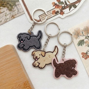 May include: Three dog-shaped keychains in gray, tan, and brown, each with a silver keyring and chain. The keychains are embroidered with a playful design, featuring a smiling dog with its tongue out. The brown keychain has a pink border.