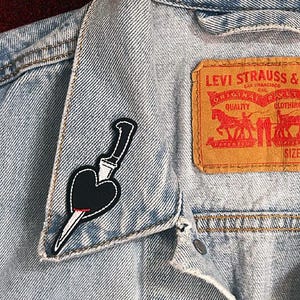 May include: Light blue denim jacket with a black heart-shaped patch pierced by a dagger. The jacket features a Levi Strauss & Co. label. The patch is on the collar of the jacket.
