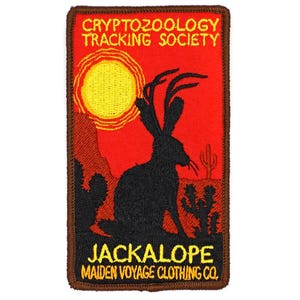 May include: Embroidered patch featuring a black jackalope silhouette against a red desert landscape with a yellow sun. Text includes "CRYPTOZOOLOGY TRACKING SOCIETY" and "JACKALOPE MAIDEN VOYAGE CLOTHING CO." with a brown border.