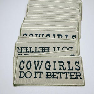 May include: A stack of rectangular patches with the text "COWGIRLS DO IT BETTER" embroidered in dark green thread on a light tan background. The patches have a light green border.