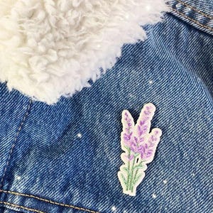 May include: A denim jacket with a white, fluffy collar and a lavender flower patch. The patch features purple flowers with green stems and leaves, adding a touch of nature to the blue jacket.