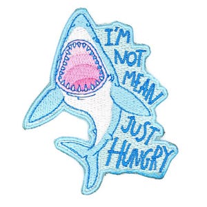 May include: Embroidered patch featuring a cartoon shark with an open mouth, displaying pink gums and white teeth. The shark is light blue with the text "I'M NOT MEAN JUST HUNGRY" in blue.