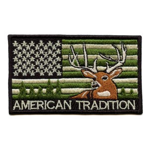 May include: Embroidered patch featuring a deer superimposed on an American flag design. The flag incorporates green and white stripes, with a black border and the text "AMERICAN TRADITION" at the bottom. The deer is brown with large antlers.