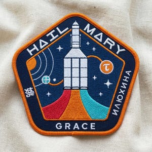 May include: A navy blue embroidered patch with an orange border. The patch features a rocket ship design with the words "HAIL MARY" and "GRACE". Additional text in a foreign language is also present.