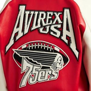 Puede incluir: Chaqueta universitaria roja y blanca con el texto "AVIREX US A" en blanco. Debajo, un gr&aacute;fico de f&uacute;tbol con un perfil nativo americano y el texto "75ers". La chaqueta tiene un cuerpo rojo y mangas blancas.