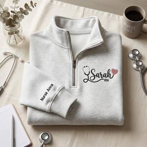 May include: A light gray quarter-zip sweatshirt featuring black embroidered text that reads Sarah RN with a stethoscope graphic and a small pink heart. The sleeve is embroidered with the text Nurse Anna in black thread on the cuff area.