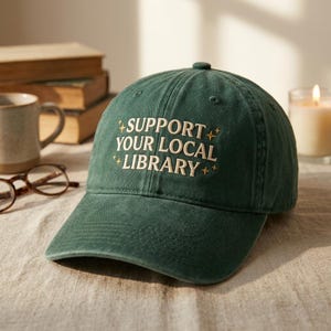 May include: A green baseball cap with the embroidered text "SUPPORT YOUR LOCAL LIBRARY" in white. The cap has a curved brim and a classic six-panel design. The text is surrounded by star-like embellishments.