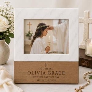 May include: A rectangular photo frame with a white marble top and a brown wood base. The wood base is engraved with a cross, the text "GOD BLESS OLIVIA GRACE ON YOUR BAPTISM DAY AUGUST 12, 2026," and holds a photo of a child receiving a baptism.