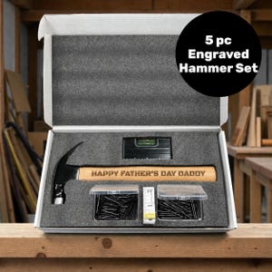 May include: A 5-piece engraved hammer set in a white box with grey foam inserts. The set includes a hammer with the text HAPPY FATHER'S DAY DADDY on the wooden handle, a black level, two clear plastic containers of nails, and a white measuring tape.