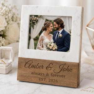 May include: A rectangular photo frame featuring a white marble top section and a dark wood base. The wood base is engraved with the text Amber & John, always & forever, and EST. 2026. The frame holds a photograph of a couple in wedding attire.
