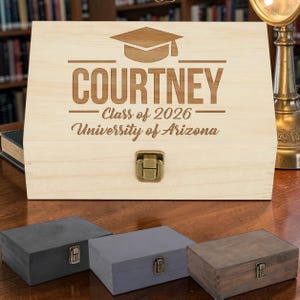 May include: A light-colored wooden box with a graduation cap design and the name "COURTNEY" engraved on the front. Below the name are the words "Class of 2026" and "University of Arizona". Three smaller boxes are in the foreground.
