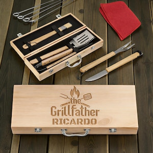 May include: A wooden barbecue set with tools, including a spatula, brush, and tongs, presented in a wooden case. The case is engraved with "the Grillfather" and a name. Additional skewers, a fork, knife, and red oven mitt are also included.