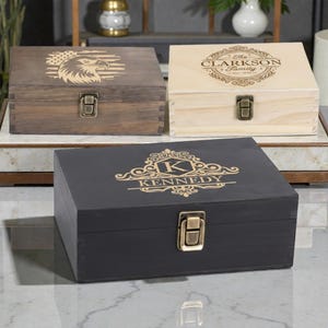 May include: Three wooden boxes with metal clasps. One box is dark brown with an eagle and American flag design. Another is light wood with "The Clarkson Family" text. The third is black with "Kennedy" text.
