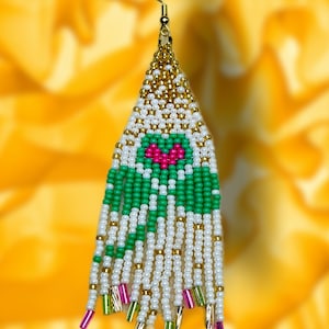 May include: Handmade beaded earring with a gold-colored hook. The earring features a triangular design with white, green, and gold beads. A pink heart shape is in the center. The bottom has dangling strands with pink and green accents.