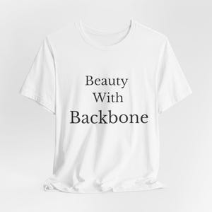 May include: A white t-shirt featuring the text "Beauty With Backbone" in black. The shirt has a crew neck and short sleeves. The material looks like cotton.