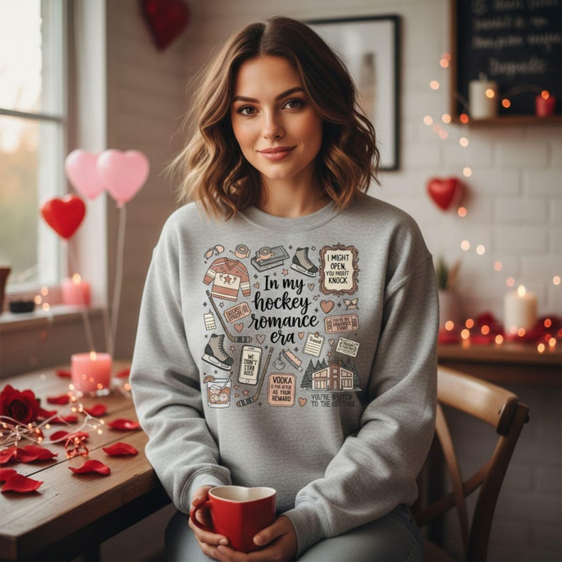 K&ouml;nnte beinhalten: Ein hellgraues Sweatshirt mit dem Aufdruck "In my hockey romance era" und verschiedenen Hockey-Grafiken. Das Design umfasst ein Hockey-Trikot, Schlittschuhe und den Text "I might open you might knock". Die Person h&auml;lt eine rote Tasse.