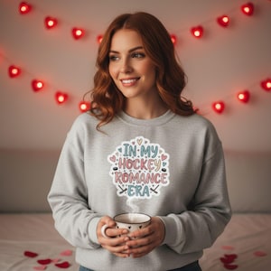 K&ouml;nnte beinhalten: Ein hellgraues Sweatshirt mit dem Text "IN MY HOCKEY ROMANCE ERA" in einem farbenfrohen Design. Das Design umfasst Herzen, Hockeyschl&auml;ger und Pucks. Die Person h&auml;lt eine wei&szlig;e Tasse mit einem schwarzen Herzmuster.