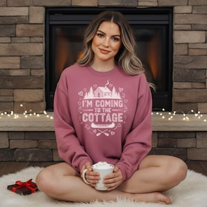 K&ouml;nnte beinhalten: Ein mauvefarbenes Sweatshirt mit dem wei&szlig;en Text "I'M COMING TO THE COTTAGE". Das Design umfasst eine H&uuml;tte, B&auml;ume, Herzen, ein Kanu und gekreuzte Paddel. Das Sweatshirt wird von einer Person getragen.