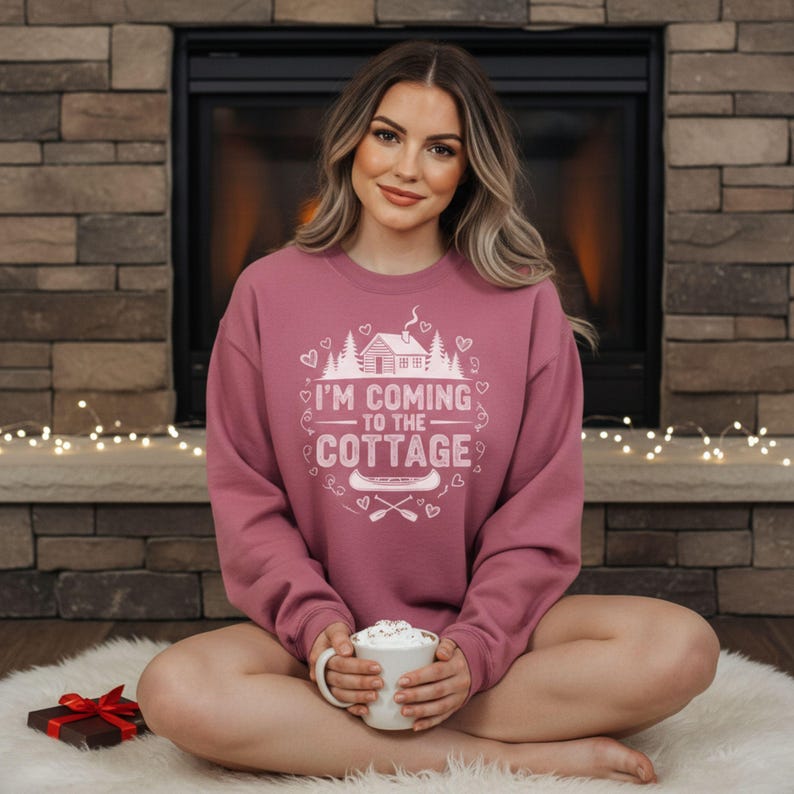 K&ouml;nnte beinhalten: Ein mauvefarbenes Sweatshirt mit dem wei&szlig;en Text "I'M COMING TO THE COTTAGE". Das Design umfasst eine H&uuml;tte, B&auml;ume, Herzen, ein Kanu und gekreuzte Paddel. Das Sweatshirt wird von einer Person getragen.