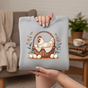 May include: A light gray sweatshirt featuring a cartoon hen sitting in a woven basket with eggs. The hen has a red comb and is surrounded by floral and leaf accents. Several eggs are placed around the basket.