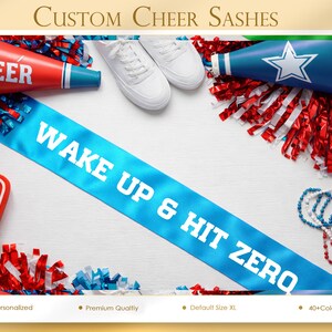 May include: A light blue sash with the text "WAKE UP & HIT ZERO" is displayed diagonally across the image. Cheerleading accessories, including red and blue pom-poms, megaphones, and white trainers, are arranged around the sash. The words "Custom Cheer Sashes" are at the top.