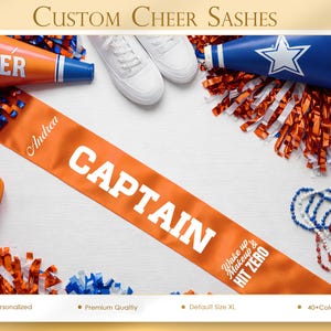 May include: An orange cheerleading sash with the word "CAPTAIN" in white. The sash also has the name "Andrea" and the text "Wake up & HIT ZERO". Cheerleading accessories, including pom-poms and a megaphone, are arranged around the sash.