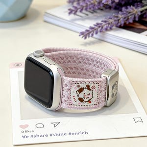 Front view of a pastel pink knit Apple watch band featuring a breathable hollow-out texture, an engraved metal slider, and a cute kawaii animal illustration patch, isolated on a white background.