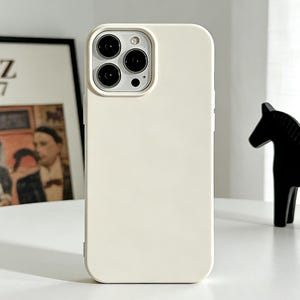 Puede incluir: Funda de teléfono de color crema con un recorte para la cámara. La funda se muestra frente a un póster enmarcado "Jazz 1947" y una figura de caballo negro. La funda tiene un acabado liso y mate.