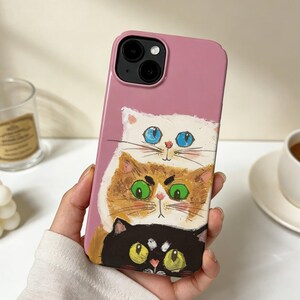 May include: A pink phone case with a quirky design of three cats, one on top of the other. The top cat is white with blue eyes, the middle cat is ginger with green eyes, and the bottom cat is black with yellow eyes. The case has a shiny finish.