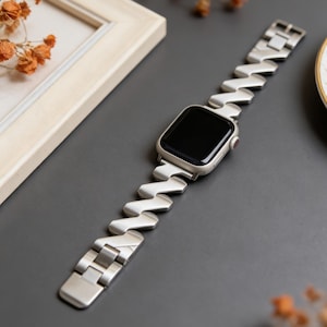 May include: A silver smartwatch with a unique zigzag-patterned band. The watch face is black, and the band is made of a shiny, metallic material. The watch is displayed on a dark gray surface.