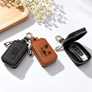 May include: Three rectangular key cases with silver zippers and metal clips. One black case features a letter A, one tan case shows a palm tree graphic with the name Harrison, and one open black case contains a black and silver car key fob.