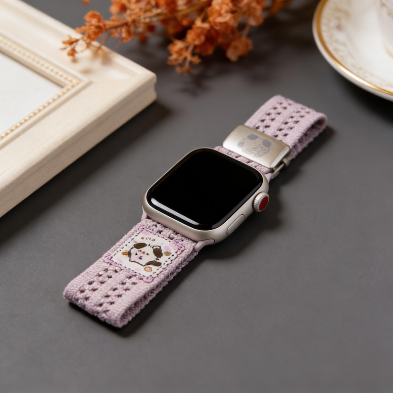 May include: A silver smartwatch with a lavender fabric band featuring a decorative dog design. The band has a metal clasp and a perforated pattern. The watch is displayed on a gray surface.