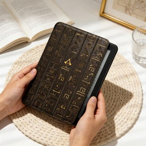 Könnte beinhalten: Eine braune E-Reader-Hülle mit goldenen Hieroglyphen-Mustern und dem Assassin's Creed-Logo. Die Hülle ist geöffnet und zeigt den Bildschirm des E-Readers. Im Hintergrund befinden sich ein Buch, ein Glas Wasser und ein gerahmtes Bild.