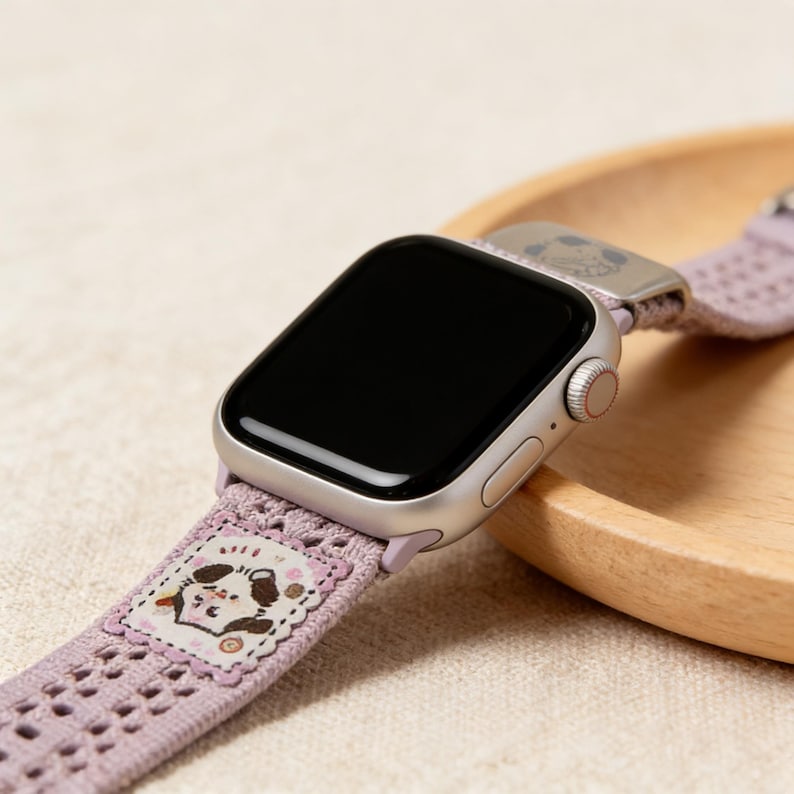 May include: Silver smartwatch with a lavender perforated band. The band features a decorative patch with a cartoon dog illustration. The watch is displayed on a wooden tray, showcasing the band's design and the watch's sleek appearance.