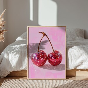 Puede incluir: Impresión enmarcada de dos cerezas con efecto bola de discoteca. Las cerezas rojas tienen detalles brillantes sobre un fondo rosa. La impresión está en un marco dorado, adecuada para la decoración de paredes.