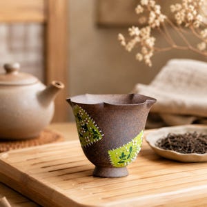 Könnte beinhalten: Eine braune Keramik-Teetasse mit einem einzigartigen, wellenförmigen Rand und einem dekorativen grün-weißen Blumenmuster. Die Teetasse steht auf einem Holztablett, im Hintergrund eine Teekanne und eine kleine Schale mit Teeblättern. Ein schönes Stück für Teeliebhaber.
