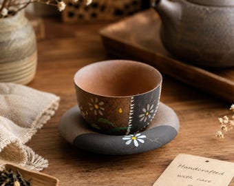 May include: A ceramic tea cup and matching saucer featuring a two-tone brown and black finish with hand-painted white daisy motifs. The cup has a smooth interior and a textured exterior, while the saucer displays a single white daisy.