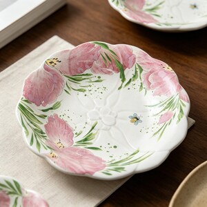 Può includere: Piatto in ceramica bianca con bordo smerlato, decorato con motivi floreali rosa e foglie verdi. Sul piatto è dipinta una piccola ape in stile cartone animato. Il piatto è su una superficie di lino beige.