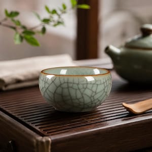 May include: A small, round tea cup featuring a pale celadon green glaze with a prominent dark crackle pattern throughout the exterior surface. The rim of the cup is finished with a thin, smooth brown band, providing a contrast to the textured body.