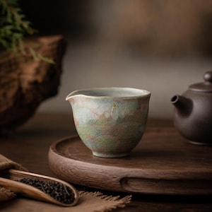 May include: A small, celadon-colored ceramic tea cup with a spout, featuring a crackle glaze. The cup sits on a wooden tray, with a dark brown teapot in the background. A wooden spoon with tea leaves is in the foreground, suggesting a tea ceremony.