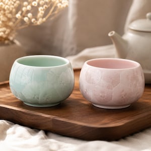 May include: Two small, round ceramic teacups, one mint green and one pale pink, sit on a wooden tray. The cups have a textured, crystalline pattern. A white teapot is visible in the background, suggesting a tea set.