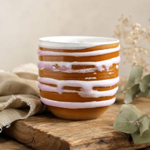 May include: A handcrafted ceramic mug with a unique design. The mug features horizontal bands of brown, white, and pink glaze. The white glaze drips down the sides, adding a textured effect. The mug sits on a wooden surface.
