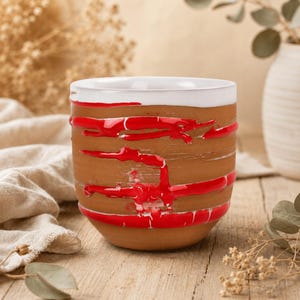 May include: A handmade ceramic mug with a brown exterior and a white interior rim. The mug is adorned with horizontal red paint strokes. The mug sits on a wooden surface, suggesting a rustic or artisanal theme.