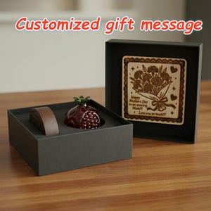 May include: A gift set featuring a decorative pomegranate figurine with a green top and red body, a wooden plaque with a floral design, and a brown object, all presented in a black box. The plaque reads "Happy Mothers Day to an amazing Mom!!! Love you so much!!!".
