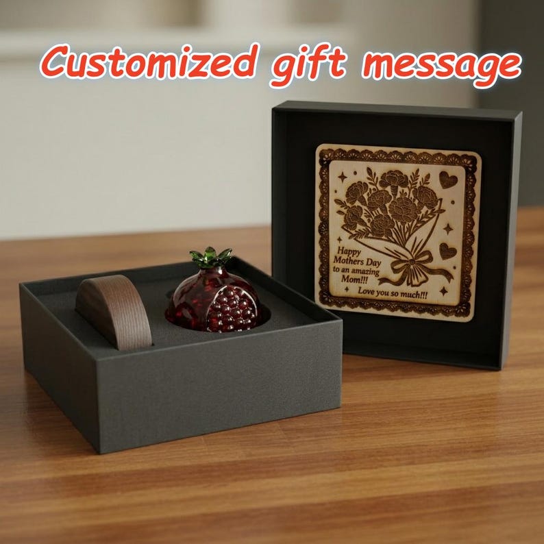 May include: A gift set featuring a decorative pomegranate figurine with a green top and red body, a wooden plaque with a floral design, and a brown object, all presented in a black box. The plaque reads "Happy Mothers Day to an amazing Mom!!! Love you so much!!!".